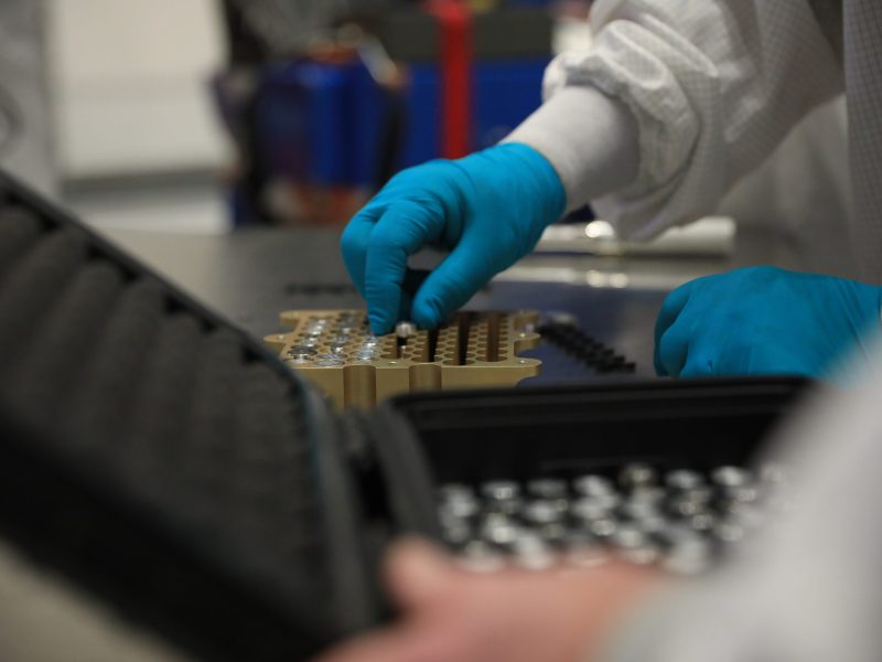 A photo of a technician's hand placing small, sealed vials into a metal array for integration into a spacecraft.