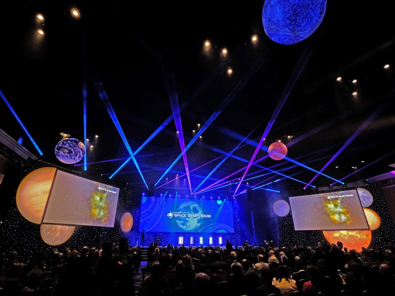 Photo of a crowd looking up at a stage decorated with lights, hanging globes, and laser effects. Multiple screens display space imagery with the words "Space Symposium."