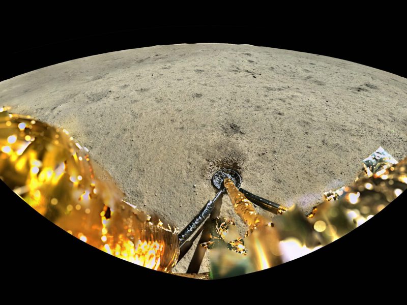 A view from the Chang'e-6 lander, showing the golden body of the spacecraft in the foreground, the impression of a landing leg in the lunar regolith, a relatively smooth lunar surface, and distant peaks and rim of Apollo crater against a black backdrop.