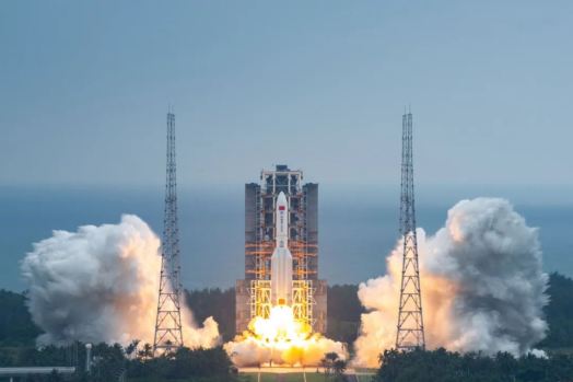 Orange exhaust pushes a large Long March rocket from the pad at the coastal Wenchang spaceport, with white smoke billowing around lightning towers near the launch tower.