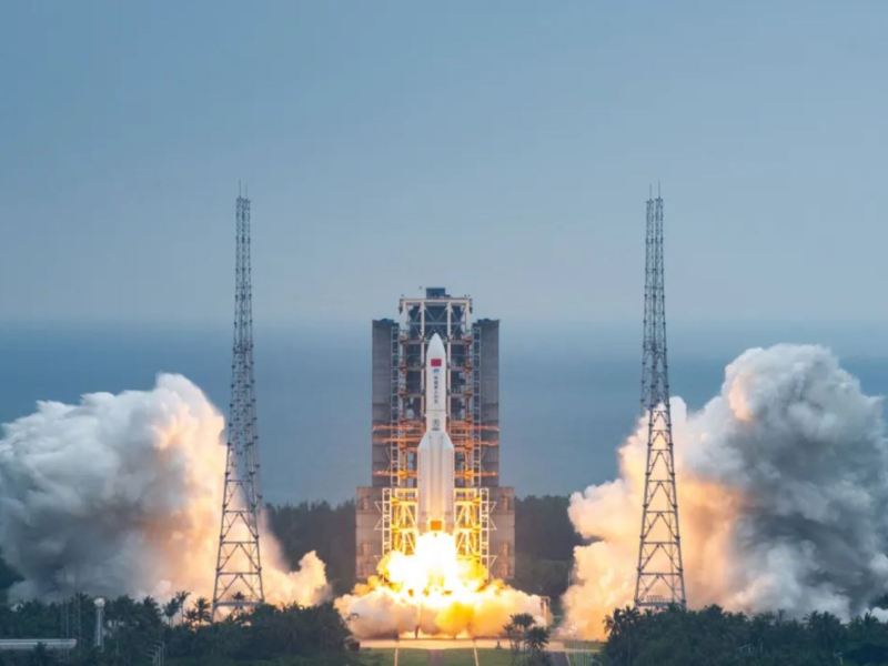 Orange exhaust pushes a large Long March rocket from the pad at the coastal Wenchang spaceport, with white smoke billowing around lightning towers near the launch tower.