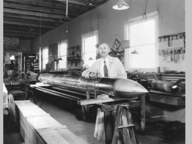 Black and White photo of Robert Goddard standing next to a rocket in a workshop.