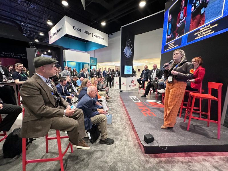 A woman in brown leather Star Wars jacket and orange pants speaks to a crowd gathered at a trade show exhibit for space company Redwire.