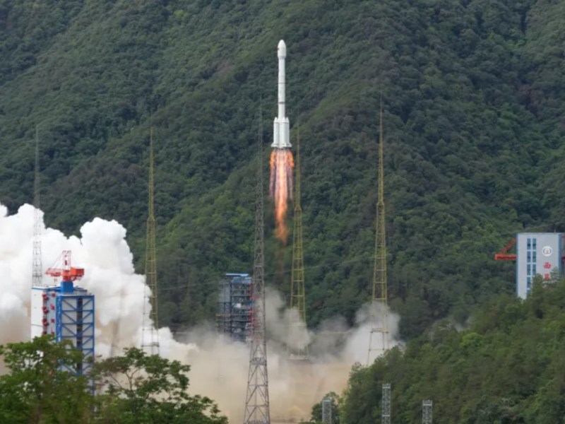 A Long March 3B rocket clears the tower above Xichang spaceport and white clouds of exhaust, amid a background of verdant hills.